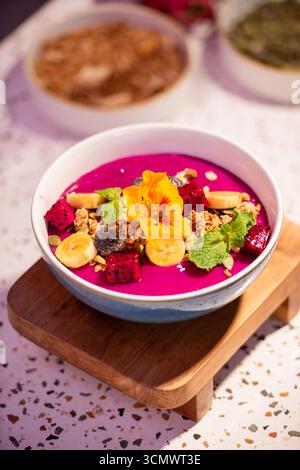 Farbenfrohe Drachenfrucht-Smoothie-Schüssel mit Banane, Müsli, essbarer Blume und frischen Minzblättern. Gesundes Frühstückskonzept mit lebhaften tropischen Früchten Stockfoto