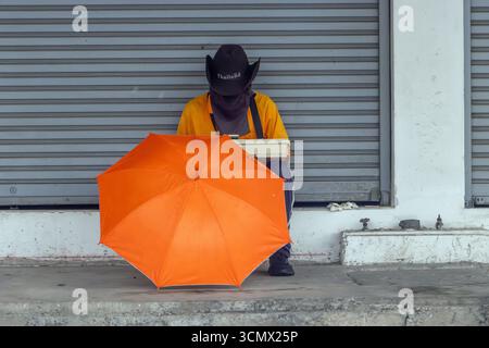 Ein maskierter Verkäufer, der einen schwarzen Hut trägt, sitzt einsam hinter einem leuchtenden orangen Regenschirm auf einer Straße in Bangkok, Thailand. Stockfoto