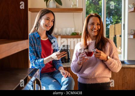 Asiatische Mutter und Tochter chatten während sie Tee trinken und Smartphone in der Küche überprüfen Stockfoto