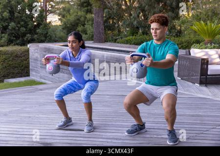 Verschiedene Freunde halten blaue und pinkfarbene Kettlebells und hocken auf der Terrasse neben dem erhöhten Pool Stockfoto