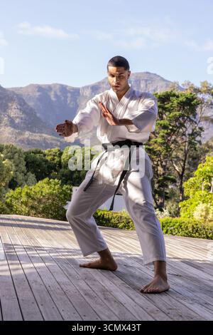 Ein Mann, der auf Holzdeck in den Bergen mit Gi und Gürtel eine niedrige Karate-Haltung übt Stockfoto