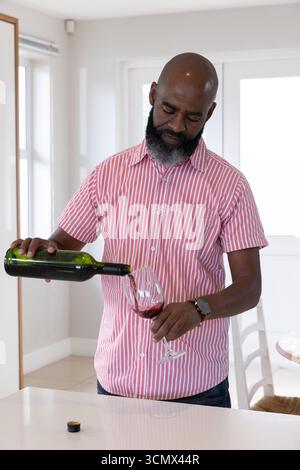 Afroamerikanischer Senior-Mann, der Rotwein aus grüner Flasche in Glas an der weißen Küchentheke gießt Stockfoto