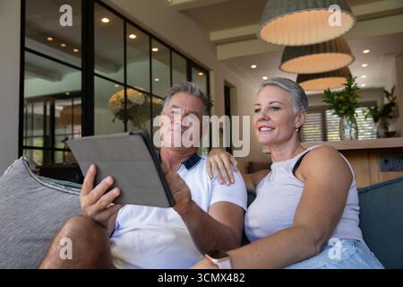 Seniorenpaar, das auf einem gepolsterten Sofa in einem modernen Wohnzimmer sitzt und ein Tablet in der Nähe der Bar verwendet Stockfoto