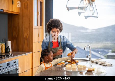 afroamerikanische Mutter und Tochter dekorieren Cupcakes auf der Kücheninsel mit Spachtel Stockfoto