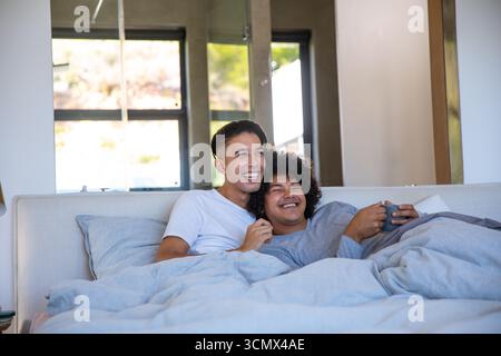 Zwei Männer kuscheln sich im hellen Schlafzimmer unter grauer Bettdecke auf dem Bett und halten den Becher unter dem Sonnenlicht Stockfoto