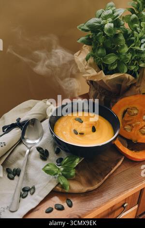 Stillleben mit heißer Kürbissuppe in einer Schüssel, halbiertem Kürbis, Löffel, Kürbiskernen und Stoff auf einem rustikalen Holztisch. Stockfoto