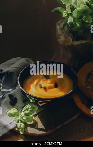 Stillleben mit heißer Kürbissuppe in einer Schüssel, halbiertem Kürbis, Löffel, Kürbiskernen und Stoff auf einem rustikalen Holztisch. Stockfoto