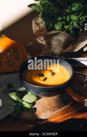 Das Leben von Kürbissuppe in einer Schüssel, halbiertem Kürbis, Löffel, Kürbiskernen und Stoff auf einem rustikalen Holztisch, beleuchtet durch warmes Sonnenlicht Stockfoto