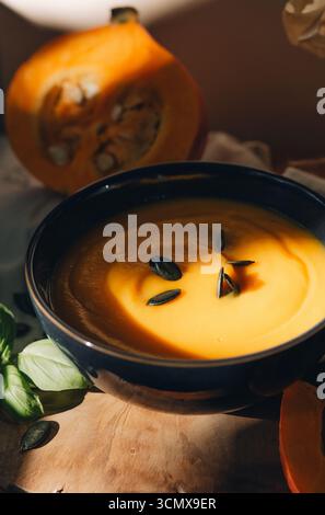 Das Leben von Kürbissuppe in einer Schüssel, halbiertem Kürbis, Löffel, Kürbiskernen und Stoff auf einem rustikalen Holztisch, beleuchtet durch warmes Sonnenlicht Stockfoto
