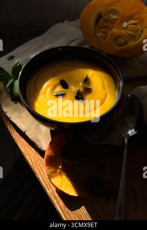 Das Leben von Kürbissuppe in einer Schüssel, halbiertem Kürbis, Löffel, Kürbiskernen und Stoff auf einem rustikalen Holztisch, beleuchtet durch warmes Sonnenlicht Stockfoto