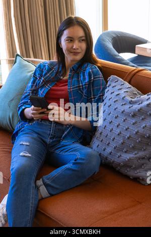 Asiatische Frau, die auf einem orangefarbenen Sofa sitzt und ein Smartphone im Wohnzimmer mit Kissen und Vorhang hält Stockfoto