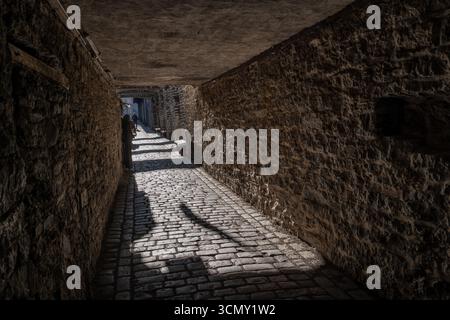 Sonnendurchfluteter Durchgang unter dem Gebäude mit schmaler Kopfsteinpflasterstraße, Steinmauern und niedriger Decke in der mittelalterlichen Altstadt in Tallinn, Estland. Stockfoto