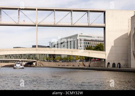 Berlin, Deutschland - 1. September 2025: Das Sonnenlicht reflektiert vom Fluss, während ein Boot an zeitgenössischen Gebäuden in Berlin vorbeifährt. Diese Szene erfasst die Stockfoto