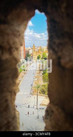 Blick durch eine Steinmauer in Richtung Serranos-Brücke und historische Gebäude in Valencia, Spanien, mit Leuten, die die Straße entlang laufen. Stockfoto