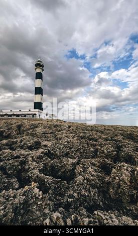 Hoher schwarz-weißer Leuchtturm am felsigen Ufer unter dramatischem bewölktem Himmel. Insel Menorca Stockfoto