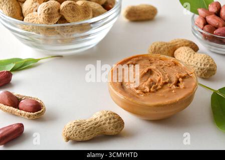 Detail von Erdnussbutter in einem Glasgefäß mit einem Metall- und Holzspachtel und einer vollen Schüssel und geschälten und geöffneten Erdnüssen, die Körner mit Blättern auf einem wh zeigen Stockfoto