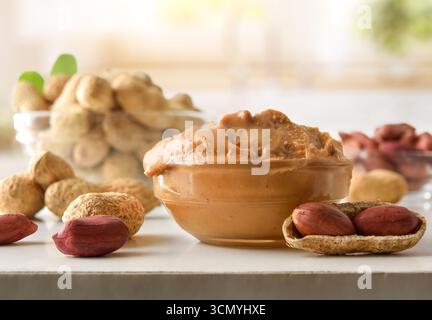 Detail einer Glasschale gefüllt mit Erdnussbutter auf einer weißen Küchenbank mit Obst und einer hellen Küche im Hintergrund. Vorderansicht. Stockfoto