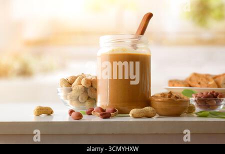 Glas und Schüssel gefüllt mit Erdnussbutter auf einem weißen Tisch mit Schalenfrüchten und ungeschälten Nüssen und Brotscheiben in einer hellen Küche. Vorderansicht. Stockfoto