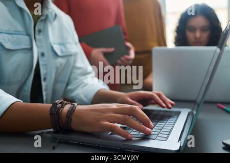 Junge Erwachsene kaukasische Frau, die auf einem Laptop mit einer multiethnischen Gruppe von Teenagern schreibt, die im Hintergrund zusammenarbeiten und sich auf ein Gemeinschaftsprojekt oder eine Lernsitzung in modernem Ambiente konzentriert Stockfoto