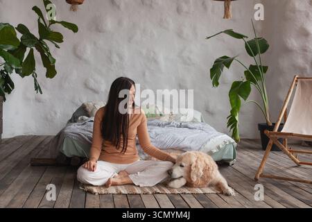 Eine Frau sitzt auf einem Teppich in einem ruhigen Hotelzimmer und übt neben ihrem freundlichen Golden Retriever Achtsamkeit und Yoga. Der Raum ist mit Pflanzen gefüllt Stockfoto