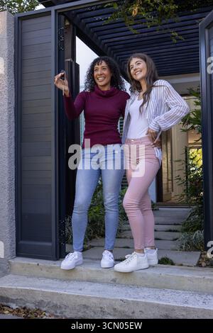 Verschiedene Freundinnen halten Schlüssel hoch und feiern neues Zuhause unter Pergola am Hauseingang. Feier, Freundschaft, Hausbesitz, Modernarchit Stockfoto