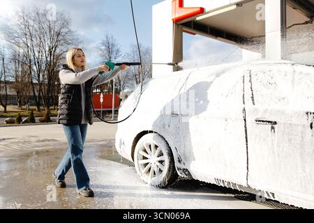 Eine junge Frau, die ihr Auto in einer Autowaschanlage wäscht und an einem sonnigen Tag Schmutz und Schmutz mit einem Hochdruckreiniger entfernt Stockfoto