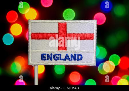Die mit England aufgestickte Flagge mit dem Heiligen Georgskreuz und Text, die vor den bokeh-Lichtern in Regenbogenform steht, die an ein Feuerwerk und eine Weihnachtsfeier erinnern. Stockfoto