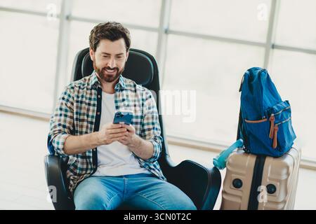 Lächelnder Mann am Flughafen, der ein Smartphone benutzt, das neben dem Gepäck liegt, bereit für eine Reise während der Ferien Stockfoto