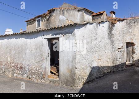 Die Fassade eines zerbröckelnden, verlassenen Hauses mit einer kaputten Tür und verwitterten Wänden, die auf den Verlauf der Zeit in einem ruhigen portugiesischen Dorf hinweisen. Stockfoto