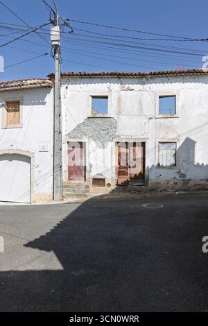 Ein verlassenes portugiesisches Haus mit fehlenden Fenstern und einer kaputten, verwitterten Tür steht auf einer sonnendurchfluteten Straße, deren verblasste Wände eine Geschichte erzählen Stockfoto