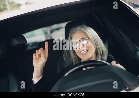 Elegante Geschäftsfrau, die ihre Fahrt an einem sonnigen Tag genießt und in einer urbanen Umgebung tatkräftig hinter dem Steuer lächelt Stockfoto