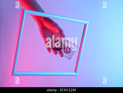 Kreative Kunst. Weibliche Hand, die die Audiokassette durch einen hochragenden Rahmen mit holografischem Neonlicht hält. Verlaufsglühen. Stockfoto