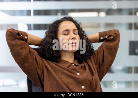 Lockige Frau lehnt sich mit den Händen hinter dem Kopf zurück, die Augen geschlossen, und genießt eine friedliche achtsame Pause an ihrem modernen Schreibtisch, ruhig und zufrieden während der Arbeit Stockfoto