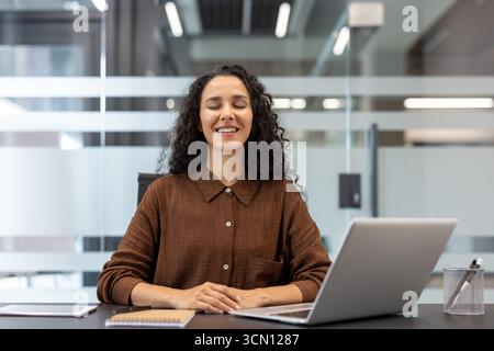 Frau mit geschlossenen Augen und einem ruhigen Ausdruck, die mit einem Laptop am Schreibtisch sitzt und während eines konzentrierten Meditationsmoments auf der Arbeit Ruhe findet und Stress reduziert Stockfoto