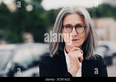Reife, reife Geschäftsfrau mit grauen Haaren in formeller Kleidung posiert an einem geschäftigen Tag in einer Stadtstraße Stockfoto