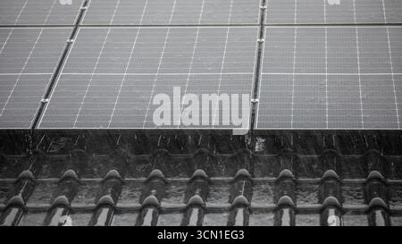 Schwarze Solarpaneele auf glänzend schwarzen Dachziegeln bei starkem Regen, tropfenförmige Tropfen auf Paneelen, Nahansicht der Dachfläche, Format 16:9 Stockfoto