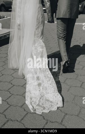 Nahaufnahme einer Braut in Spitzenkleid und Schleier, die beim Gehen Hände mit dem Bräutigam hält, Schwarz-weiß-Bild, elegante Hochzeitsmode und romantische Atmosphäre. Stockfoto
