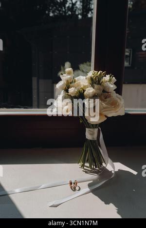 Eleganter Blumenstrauß aus weißen Rosen mit Satinband und Eheringen, die auf einer Fensterbank angeordnet sind und in natürlichem Licht für eine romantische Hochzeitsatmosphäre eingefangen werden Stockfoto