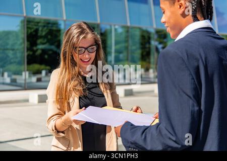 Multikulturelle Geschäftspartner, die Dokumente außerhalb eines modernen Bürogebäudes prüfen, diskutieren ein Projekt, das Teamarbeit und Zusammenarbeit darstellt Stockfoto