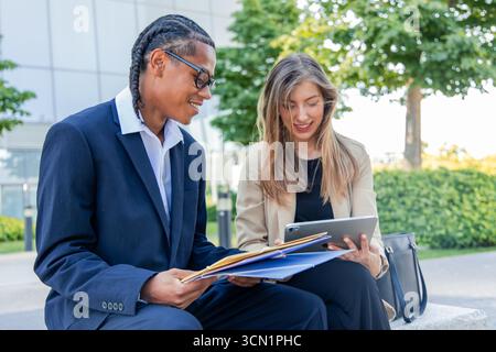Multiethnische Geschäftspartner, ein junger Mann und eine junge Frau, sitzen draußen im Finanzviertel, prüfen Dokumente und verwenden ein Tablet, um zu arbeiten Stockfoto