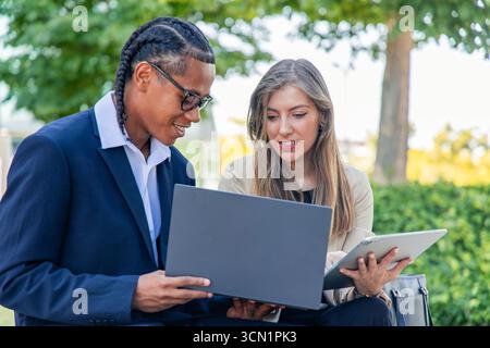 Geschäftspartner arbeiten an einem Projekt zusammen, verwenden Laptop und Tablet, diskutieren Strategien und teilen Ideen in einem Park, zeigen Teamwork und Modus Stockfoto