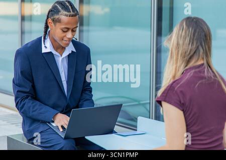 Ein junger Geschäftsmann, der mit seiner Kollegin zusammenarbeitet, während er draußen im Finanzviertel einen Laptop benutzte, saß beide an einem modernen Schreibtisch Stockfoto