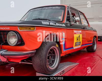 Red & Black BMW Race Car am Paddock – Ein klassischer BMW 2002 Rallyefahrer in Rot und Schwarz mit „BMW ALPINA“ auf der Motorhaube steht auf Rampen auf einer roten Plane bei einem m Stockfoto