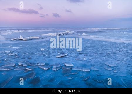 Gefrorenes Meer mit Eisrissen und gestapelten Platten unter einem pastellrosa und blauen Sonnenuntergangshimmel in winterlicher Stille. Stockfoto
