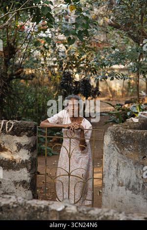 Goa, Indien - 21. September 2014: Porträt einer älteren Indianerin, die ein Sommerkleid mit Blumenmotiv trägt und auf schäbigen Zaunzaun geneigt ist, während sie für ein Foto posiert. Wilder Garten im Hintergrund Stockfoto