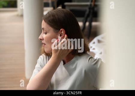 Eine Frau in einem hellgrauen T-Shirt passt ihr kurzes Haar an, zeigt silberne Creolen und rote Maniküre auf einer Terrasse Stockfoto