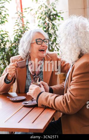 Fröhliches älteres Paar, das sich in einem herzlichen Gespräch mit Kaffee, Lächeln und Lachen einlässt und die einfachen Freuden des Lebens genießt Stockfoto