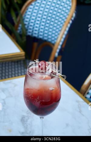 Ein wunderschöner und erfrischender roter Cocktail, der in einem Stielglas auf einem Marmortisch serviert wird. Das pulsierende rubinrote Getränk ist mit Eis und frischen Beeren gefüllt Stockfoto