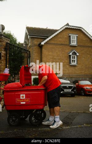 Chiswick, London, Großbritannien – 2025.08.26 – Ein Postbote von Royal Mail in roter Uniform lehnt sich über seinen Wagen, um Post auf einer Wohnstraße mit geparkten Autos zu sortieren. Stockfoto