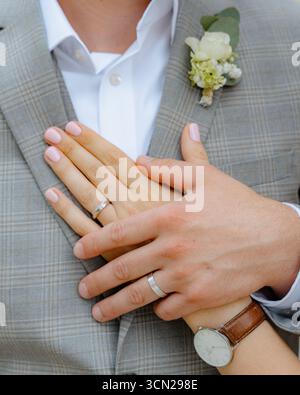 Nahaufnahme: Zwei Hände mit Eheringen, die auf einer Anzugsjacke des Bräutigams liegen. Die Frauenhand wird sanft auf die Hand des Mannes gelegt, beide tragen sie Stockfoto